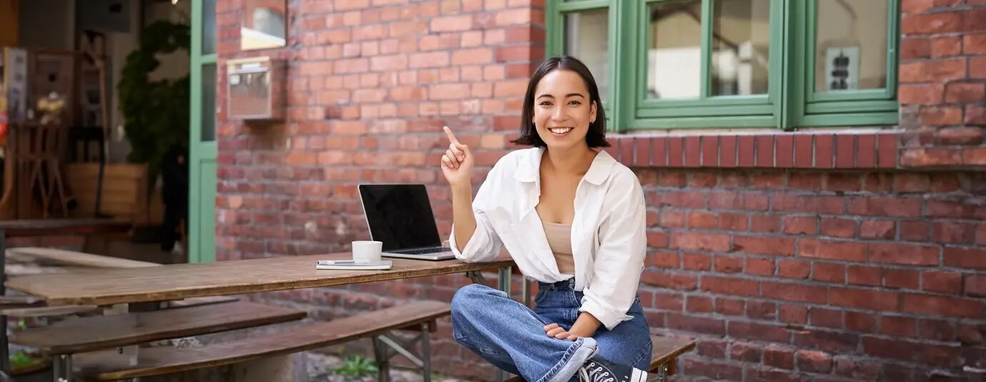 Stilbewusstes, modernes asiatisches Mädchen sitzt mit einem Laptop im Café, schaut erstaunt und zeigt nach rechts oben.