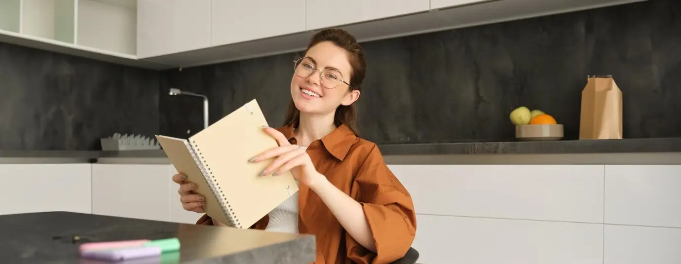Porträt einer schönen College-Studentin mit Brille, die lächelnd in der Küche mit einem Notebook sitzt.