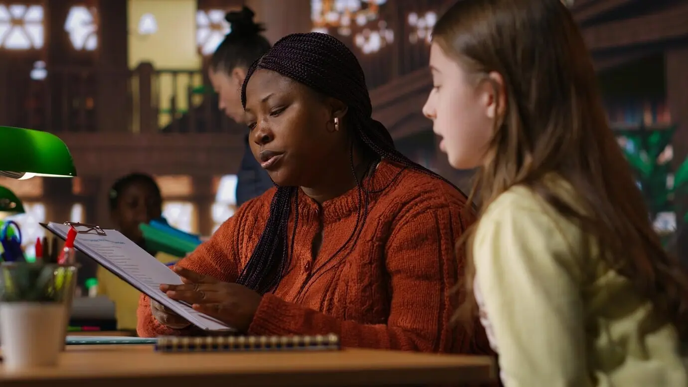 Schwarzer Nachhilfelehrer betreut eine Schülerin an einem Lerntisch in einer Schulbibliothek.